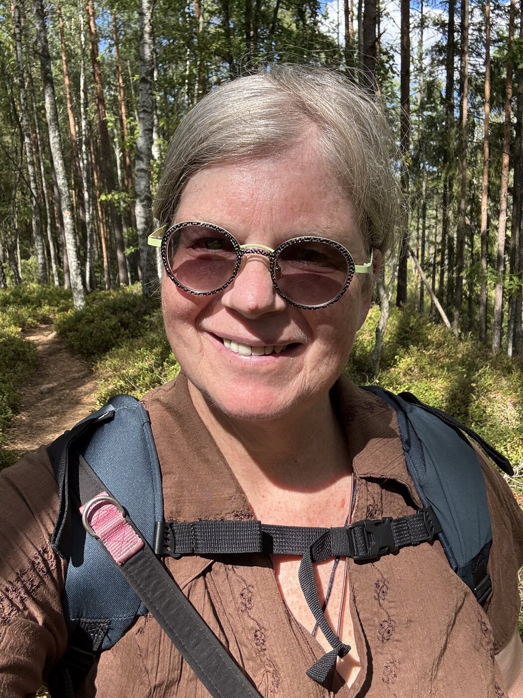Nicola lächelt mit Sonnenbrille in die Kamera, dahinter lichter Kiefernwald